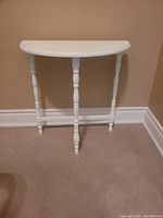White painted solid wood hall table with a semi-circular top and three turned spindle legs shown against a beige wall on carpeted floor.