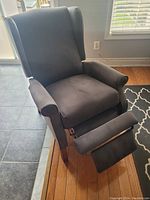 Single black fabric reclining armchair with cushioned back, armrests, and elevated footrest in a home setting with natural light.