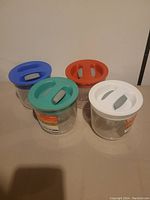Four clear round plastic storage containers with colored lids, including blue, orange, teal, and white lids. The lids have handles for easy lifting.