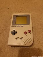 Front-facing image of the vintage Nintendo Game Boy resting on a carpeted floor, showing the screen, buttons, and speaker grille clearly.