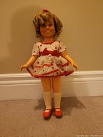 Photo of a vintage doll standing against a beige wall wearing a white dress with red polka dots and trim, red shoes, white knee socks, with molded curly blonde hair tied with a red bow.