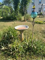 Outdoor photo of the cement bird bath and metal stake with blue-green glass ball and star in a garden setting