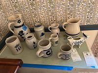 Photo of all ten pottery pieces arranged on a table showing the variety of shapes and blue-white designs, including pitchers, mugs, jars, and a ceramic bird.
