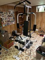 Full view of Body-Solid multi-function home gym system with padded seat and backrest, white metal frame, black padding, weight plates on the floor, in a basement room with wood paneling and various items nearby.