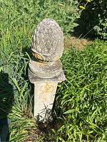 Full view of pineapple finial sculpture on a square pedestal with floral scrollwork, surrounded by garden vegetation.
