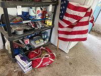 Photo shows shelving with cleaning supplies including bleach, OxiClean, Formula 409, duct tape, gloves, some tools and small spray bottles next to an American flag on a wooden pole.