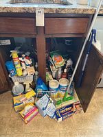 Photo showing cleaning supplies stored in and around under-sink wooden cabinet, including disinfecting wipes, sponges, spray cleaner, and mop.