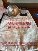 Photo showing the Federal Reserve Bank of New York money bag with red lettering, the tarnished silver bowl, and the one dollar coin set.