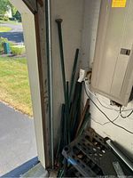 Photo showing multiple green metal stakes of varying lengths stored upright against wall near electrical panel.