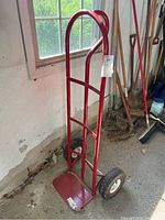 Full view of red steel hand truck upright against wall, showing handle, frame, base plate, and tires.