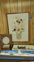 Photo shows several items: large framed needlework floral bouquet, wood box with crochet doily open on top, and several smaller framed needlework winter scenes arranged below on floor against a wood paneled wall.