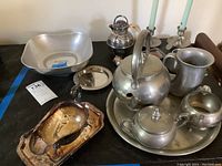 Photo shows a pewter tea set on a round tray including teapot, sugar bowl, and creamer. Also seen are two candlesticks, copper tray, silver plate tea pot with tea leaf holder, and a large pewter pitcher.