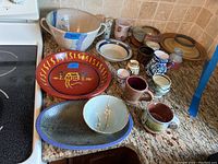 Overview of 19 pottery pieces on counter including bowls, plates, mugs, and pitchers in various colors and designs