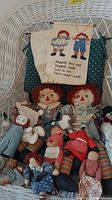Photo showing multiple Raggedy Ann and Andy dolls along with other dolls positioned in a wicker basket, includes fabric banner above them with Raggedy Ann and Andy images and poem.