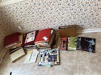 Several stacks of scrapbooks and albums alongside ballet programs and printed materials on a tiled floor with floral wallpaper background.