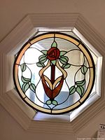 Front view of the round stained glass panel showing the floral design with red rose and green leaves framed by cream and amber glass in an octagonal shape.
