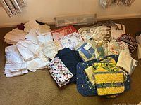 Wide view showing large assortment of vintage tablecloths, napkins, doilies, contemporary placemats, napkin rings, and pot holders spread on carpeted floor.