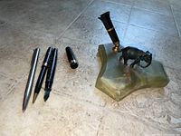 Three pens and separate pen cap with elephant stone pen holder on tiled floor.