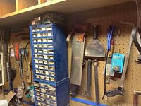 View of a tall blue metal organizer with multiple drawers, assorted hand tools including saw and putty knife hung on pegboard