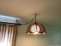 Woven hanging lamp shade illuminated and hanging from ceiling with visible curtain and ceiling molding detail in background.
