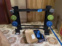Black metal hand weight stand holding 3 sets of hex dumbbells in 5 lb green, 8 lb blue, and 10 lb silver colors, and the Bollinger ankle weights and grip strengthener placed on floor in front