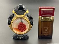 Two Cartier perfume bottles side by side on black surface, one with black glass panthers and circular shape, the other rectangular with red and gold cap.