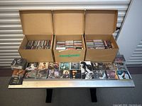 Three cardboard boxes filled with assorted music CD jewel cases, plus a row of CDs displayed in front