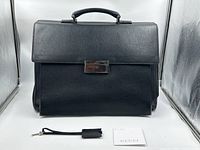 Front view of briefcase showing flap, lock, handle, key fob and original tag laid in front.