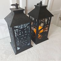 Two black metal battery operated lantern lamps, one with an owl cutout design and electric flickering candle, the other with a fox and tree cutout design and LED light.