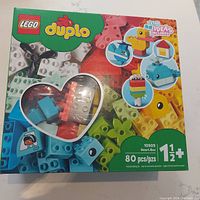 Box front showing LEGO Duplo branding, colorful blocks, assembled figure examples, and heart-shaped window revealing the blocks inside.