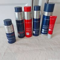 Front view of all six Clarins Men skincare products arranged in a row on a surface: 5 pump bottles in blue and silver tones plus one red tube.