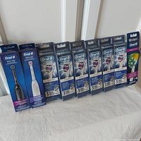 Photo showing two Oral-B Pro 100 toothbrushes (Crossaction black and 3D White white) and seven boxed packs of replacement heads (six 3D White packs and one Crossaction pack) in sealed packaging.