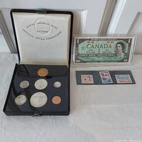Overall lot showing Royal Canadian Mint coin set in presentation box, one dollar bill in plastic case, and three stamps