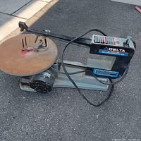 Full side view of the Delta 16 inch scroll saw showing metal base, arm, power cord, and round metal table with rust.