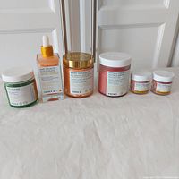Full view of six Truely Beauty products arranged in a line on a white surface: green matcha face scrub, orange anti-cellulite body serum, orange body polish, pink bronze & blur cream, two small lip products.