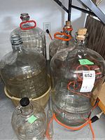 Four glass carboys grouped together, two with orange handles and two with cork stoppers. One is in a beige basket base.