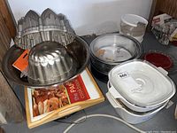 Cooking and baking items including metal cake molds, baking trays, glass trays, and mixing bowls with a cookbook beneath.