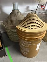 Photo showing two demijohn carboys, one beige plastic with conical plastic cover, the other a glass carboy with woven protective basket