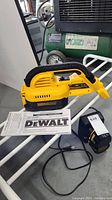 Yellow and black DeWalt cordless handheld vacuum with attached hose, battery, charger and instruction manual displayed on white metal surface.