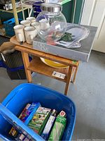 Photo showing blue tote filled with kitchen wraps and storage bags, yellow platter below wooden shelf, large glass jar with silver lid, boxed glass platter on top of wooden stand, and six white coffee mugs.