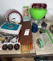 Image showing full assortment of kitchen items including wall clocks, cutlery, mixing bowls, glassware, muffin molds, and other utensils on a table.