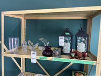 Shelf displaying Lenox glass figurines including an elephant and two dolphins, two glass insulators, a pair of metal lanterns with snowflake designs and battery-powered candles, and five small wooden cups.