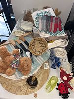 Photo showing assorted linens, stuffed toys (brown teddy bear, bunny), woven basket, wood kitchen utensils, glass and ceramic serving pieces on table