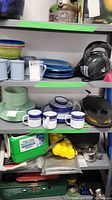 Shelves with various outdoor dishware including enamel mugs, plates, bowls, a pair of headphones, a green air pump, and picnic table set in original packaging below.