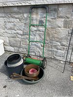 Photo showing the full manual push mower next to various plastic planters and garden trays on concrete ground by a stone wall.