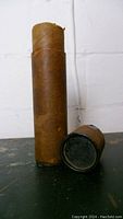 Photo of two rusted metal cylindrical containers for 60mm mortar shells from 1951, placed on table against white wall.