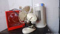 Three units on a black surface: red industrial heater, beige metal fan, and white dehumidifier.