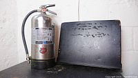 Photo showing a stainless steel fire extinguisher with hose and a black metal sheet music stand with visible wear and glue remnants on the stand surface.