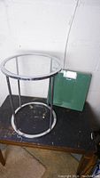 Round glass top side table with metal frame and green platform scale on a black surface, against white wall.