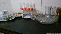 Overview of the lot showing Coke bottles, ceramic plates, and glass items on a dark surface against a white brick wall.